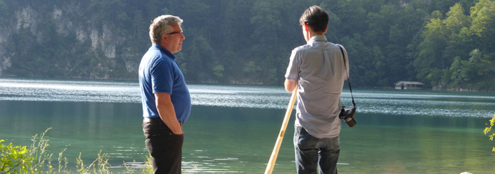 Thomas erklärt einem Freund das Alphornspiel. Im Hintergrund ist der Alpsee bei Hohenschwangau